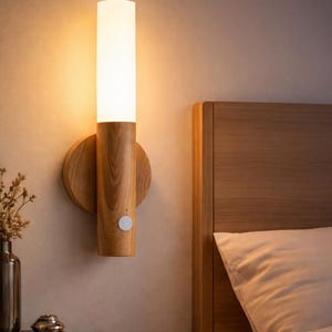 May include: A wooden wall sconce with a cylindrical frosted glass shade. The sconce is mounted on a round wooden base with a small white button. The warm light illuminates a bedroom setting, next to a wooden bed frame and nightstand.
