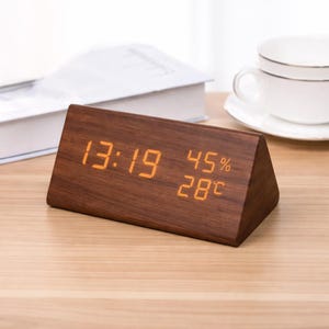 Puede incluir: Un reloj digital de madera en forma triangular, que muestra la hora, la humedad y la temperatura. El reloj tiene un acabado de madera marrón oscuro y números digitales naranjas. La hora es 13:19, con un 45% de humedad y una temperatura de 28 grados Celsius.