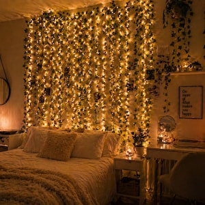 May include: A cozy bedroom scene with a bed, desk, and wall decorated with string lights and faux ivy. The bed is covered with a soft, textured blanket and pillows. A mirror and framed art add to the room's decor. The lighting creates a warm, inviting atmosphere.