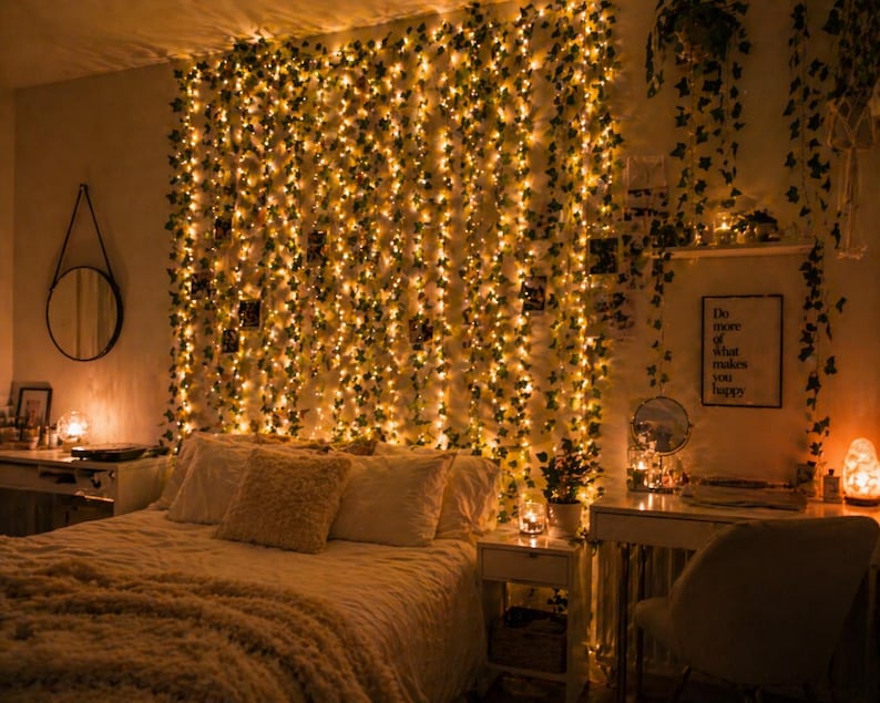 May include: A cozy bedroom scene with a bed, desk, and wall decorated with string lights and faux ivy. The bed is covered with a soft, textured blanket and pillows. A mirror and framed art add to the room's decor. The lighting creates a warm, inviting atmosphere.