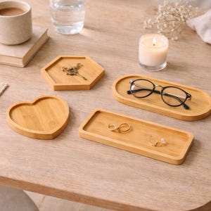 May include: A collection of bamboo trays in various shapes, including a heart, hexagon, oval, and rectangle. The trays hold items like eyeglasses, jewelry, and a decorative key. A lit candle, a cup of coffee, and a book are also on the wooden table.