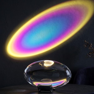 May include: A clear, oval lamp with a black base projects a rainbow-coloured light onto a dark surface. The light creates an oval shape with yellow, pink, blue, and purple hues. The lamp sits on a dark surface.