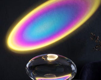 May include: A clear, oval-shaped lamp with a black base projects a vibrant, rainbow-colored light onto a dark surface. The light creates an oval shape with yellow, pink, blue, and purple hues. The lamp sits on a dark surface.