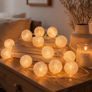 Può includere: Una catena di luci sferiche con coperture intrecciate color bianco sporco e testurizzate che emettono una luce calda e soffusa. Le luci sono disposte a grappolo, collegate da un filo sottile e trasparente per un'illuminazione accogliente.
