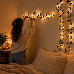 May include: A person decorates a wall with a string of fairy lights and artificial ivy. The warm glow of the lights illuminates the room, creating a cozy atmosphere. The bed is made with white pillows and a soft, textured blanket.