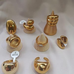 Puede incluir: Colección de anillos dorados de diversos diseños, expuestos sobre pequeños soportes de madera. Los anillos presentan formas lisas, texturizadas y geométricas. Un anillo tiene una etiqueta de talla 7. Fondo neutro.