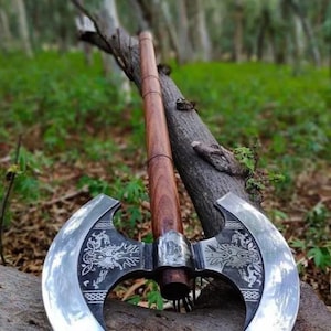May include: A double-bladed axe with a polished steel head and a wooden handle. The axe head features intricate dragon designs. The axe is leaning against a tree branch in a forest setting.
