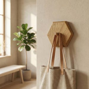 Könnte beinhalten: Ein sechseckiger Wandhaken aus Holz mit einer hellfarbenen Canvas-Tragetasche, die daran hängt. Die Tasche hat Lederriemen. Der Haken ist an einer Wand in neutralen Farbtönen befestigt. Im Hintergrund sind eine Topfpflanze und eine Holzbank zu sehen.