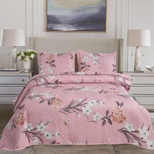 Op de afbeelding: Roze bloemenquiltset met twee bijpassende kussenslopen. Het beddengoed heeft een herhalend patroon van witte en bruine bloemen en bladeren op een gewatteerde achtergrond. Het bed is opgemaakt met de quilt en kussenslopen.