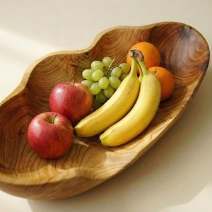 Può includere: Una fruttiera in legno piena di frutta fresca. La fruttiera contiene mele rosse, uva verde, banane mature e arance. La fruttiera ha una finitura naturale venata e una forma organica unica.