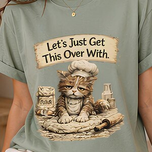 May include: Sage green t-shirt featuring a grumpy cat chef with the text "Let's Just Get This Over With." The cat is surrounded by baking supplies. The shirt is made of a soft material.
