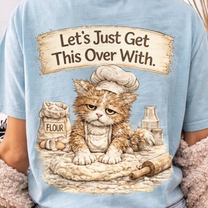 May include: Light blue t-shirt featuring a distressed graphic of a cat wearing a chef's hat, with the text "Let's Just Get This Over With." The cat is surrounded by baking supplies. A humorous design for cat lovers.