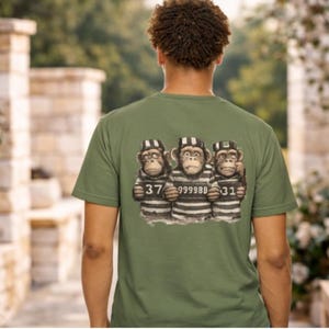 Peut inclure: T-shirt vert olive avec un motif de trois singes en uniforme de prisonnier. Chaque singe porte un numéro sur sa poitrine. Le t-shirt est porté par une personne aux cheveux bouclés.