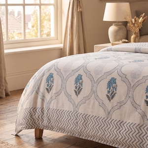 Puede incluir: Una funda nórdica blanca con un patrón floral azul repetitivo dentro de contornos festoneados, con un borde en zigzag azul y blanco en la parte inferior. La tela muestra una estética de estampado en bloque con tonos azules y grises suaves.