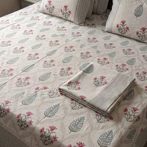 May include: A cotton bed sheet set featuring an off-white base with a repeating pattern of pink flowers and grey leaves. The set includes two matching pillowcases and a folded flat sheet, all finished with a grey chevron border design.