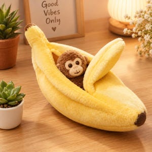 May include: A plush toy shaped like a yellow banana with a partially peeled skin, revealing a small, brown, fuzzy monkey with large eyes and a stitched smile nestled inside the center of the fruit.