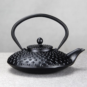 May include: Black cast iron teapot with a textured, dotted surface. The teapot has a curved handle and a spout for pouring. The lid is also black and has a small knob. The teapot is designed for brewing tea.