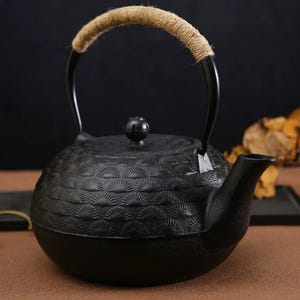 Puede incluir: Tetera de hierro fundido negro con un patrón texturizado en forma de abanico. La tetera tiene un pico curvo, una tapa con un pequeño pomo y un asa envuelta con cuerda de color natural. Diseñada para preparar té.