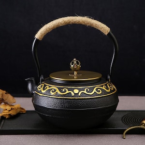 May include: Black cast iron teapot with a gold-colored lid and decorative accents. The handle is black with a tan rope grip. The teapot sits on a black tray, showcasing its traditional design.