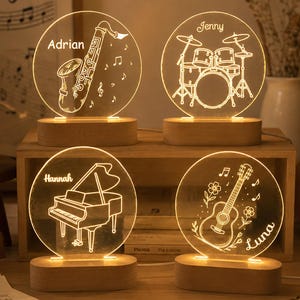 May include: Four circular acrylic night lights with warm LED illumination, each mounted on a light wood base. The designs feature engraved musical instruments: a saxophone labeled Adrian, a drum kit labeled Jenny, a piano labeled Hannah, and a guitar labeled Luna.