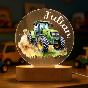 Könnte beinhalten: Personalisiertes Nachtlicht mit einem grünen Traktormotiv und dem Namen "Julian" in weißer Schrift. Der Traktor ist im Aquarellstil dargestellt, mit gelben und schwarzen Reifen. Das Licht steht auf einem Holzsockel.