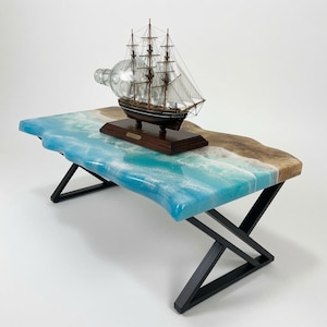 Peut inclure: Une table basse rectangulaire avec un plateau en bois à bord naturel et un design de vagues océaniques en résine bleue et blanche. La table repose sur des pieds en métal noir en forme de Z géométrique. Un navire en bouteille est posé dessus.