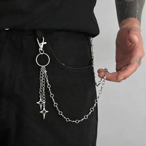 May include: Silver-toned chain with star charms attached to a belt loop. The chain is connected to a pair of black pants. The chain has a lobster clasp and a ring. The chain is held by a hand.