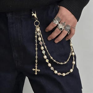 May include: Gold-tone chain wallet accessory with a cross pendant and faux pearl and gemstone accents. The chain is attached to a gold-tone clasp and is draped over dark blue denim jeans. The accessory is a unique fashion statement.