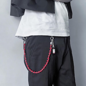 May include: A red beaded chain with silver accents and a small bottle charm attached to black pants. The chain features red beads and silver beads, with a skull charm and a small bottle with a label.
