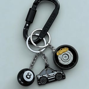 May include: Black keychain with a carabiner clip, holding a black eight ball, a car, and a brake rotor charm. The car charm has Japanese text. The brake rotor has a gold-coloured brake caliper.