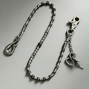 Puede incluir: Una cadena de billetera plateada con un cierre de gancho y un dije de serpiente decorativo. La cadena está hecha de cuentas negras facetadas y elementos metálicos plateados. El cierre de gancho tiene un diseño ornamentado.