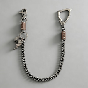 Op de afbeelding: Een zilverkleurige portemonnee ketting met een grote sluiting en een kleinere clip. De ketting is gemaakt van dikke, in elkaar grijpende schakels. Bruine lederen accenten zijn zichtbaar bij de sluitingen. Een kleine verzameling bedels hangt aan een ring bij de clip.