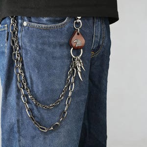 May include: A silver chain wallet chain attached to blue denim jeans. The chain has a double strand design. A brown leather tag with an eagle emblem and silver claw charms are attached to the chain.