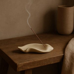 Puede incluir: Un portaincienso de cerámica beige con forma de hoja, con una varilla de incienso encendida que emite humo. El soporte está sobre una superficie de madera. En el fondo hay un jarrón texturizado. La imagen evoca paz y relajación.