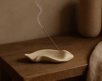 May include: A beige ceramic incense holder with a curved, leaf-like design. A lit incense stick emits a thin stream of smoke. The holder sits on a wooden surface, with a textured vase in the background. The scene evokes a sense of calm and relaxation.