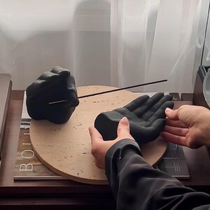 May include: A matte black ceramic incense holder shaped like a human hand, featuring a cupped palm design. A thin, dark brown incense stick is inserted into the wrist area of the hand sculpture, which rests on a circular, light beige stone base.