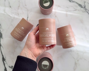 May include: A collection of cylindrical incense containers in shades of pink and brown, labeled with the scents: Sandalwood, Lavender, and Pinewood. The containers are arranged on a marble surface, with the brand name "soulscape" visible on the lid of one.