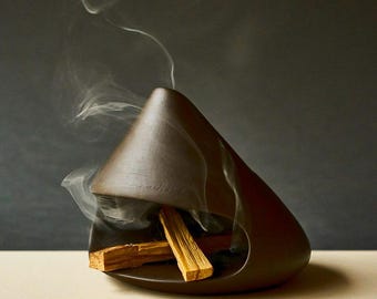 May include: A brown ceramic incense holder with a triangular shape. The holder contains several pieces of burning palo santo wood, with wisps of smoke rising from the top. The holder sits on a light-colored surface against a dark background.
