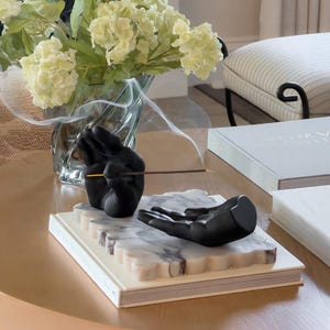 May include: A black incense holder featuring two sculpted hands resting on a rectangular white marble base with scalloped edges. One hand is positioned upright to hold a thin incense stick, while the other hand lies flat, palm facing upward.
