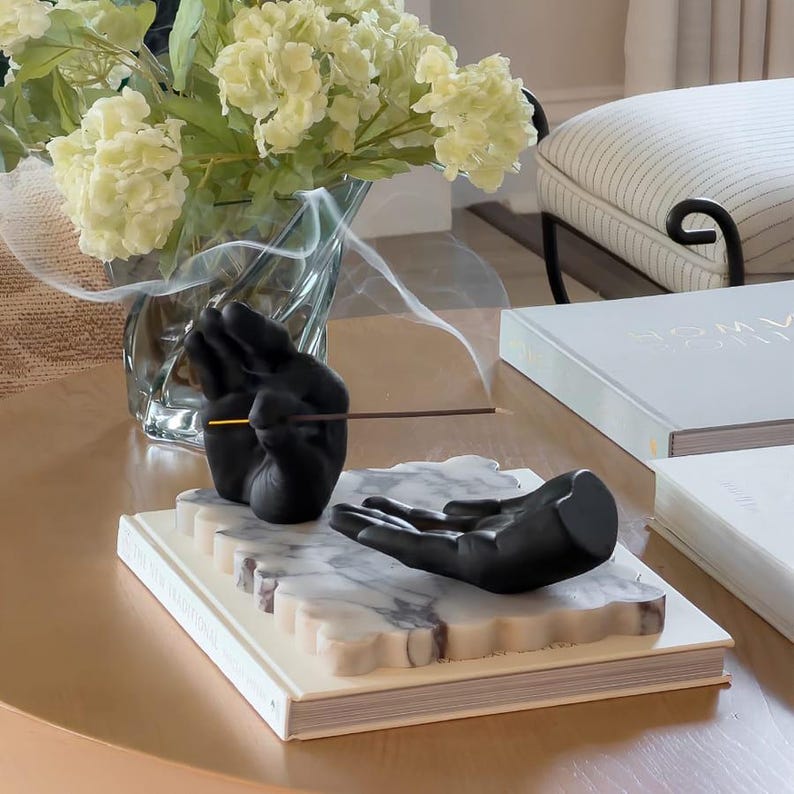 May include: A black incense holder featuring two sculpted hands resting on a rectangular white marble base with scalloped edges. One hand is positioned upright to hold a thin incense stick, while the other hand lies flat, palm facing upward.