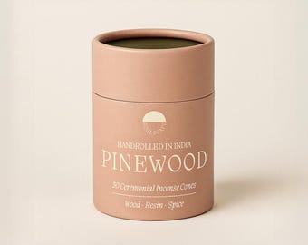 May include: A cylindrical, light pink container of incense cones. The text "HANDROLLED IN INDIA" and "PINEWOOD" are printed on the front. Below, it reads "30 Ceremonial Incense Cones" and "Wood · Resin · Spice".