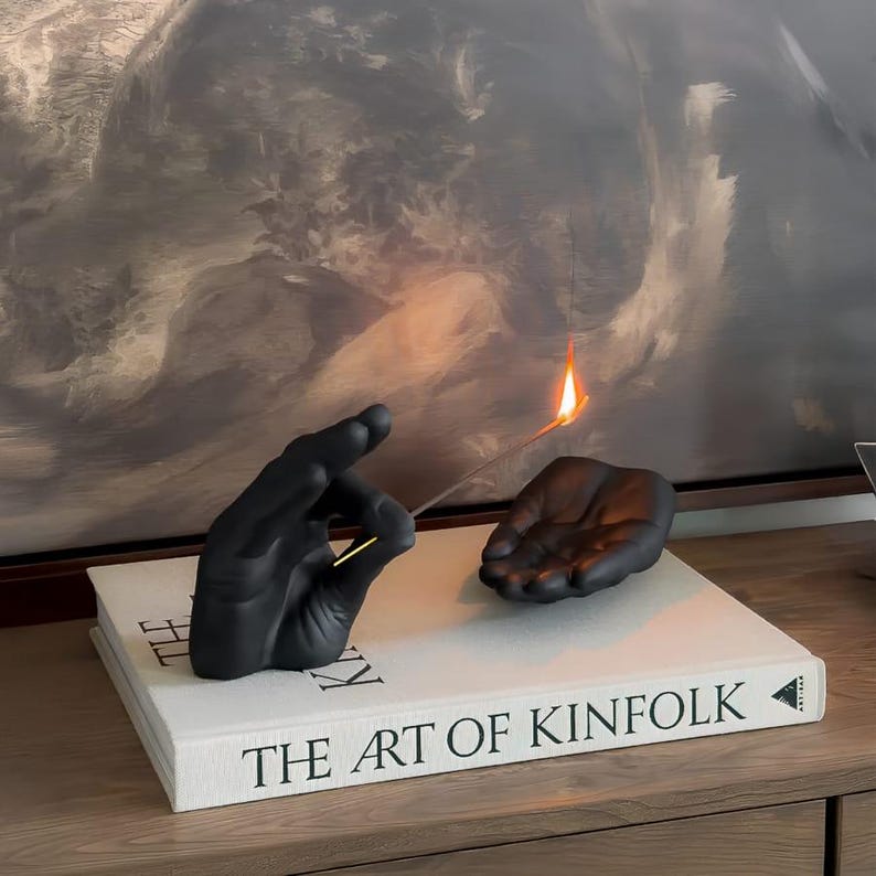 May include: Two matte black hand sculptures resting on a white book titled The Art of Kinfolk. One hand is positioned upright with fingers curled, holding a thin incense stick with a small orange flame at the tip, while the other hand lies flat.