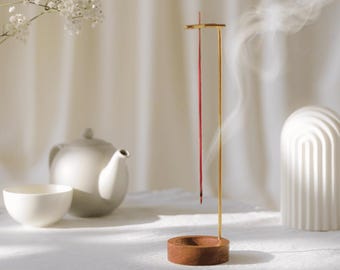 May include: A minimalist incense holder with a wooden base and a gold-colored metal stand. A red incense stick is burning, releasing a wisp of smoke. A white teapot, cup, and decorative arch are in the background.