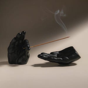 May include: A matte black incense holder set consisting of two sculpted hands. One hand is positioned vertically to hold a thin brown incense stick, while the other hand is positioned horizontally with an open palm to catch falling ash.