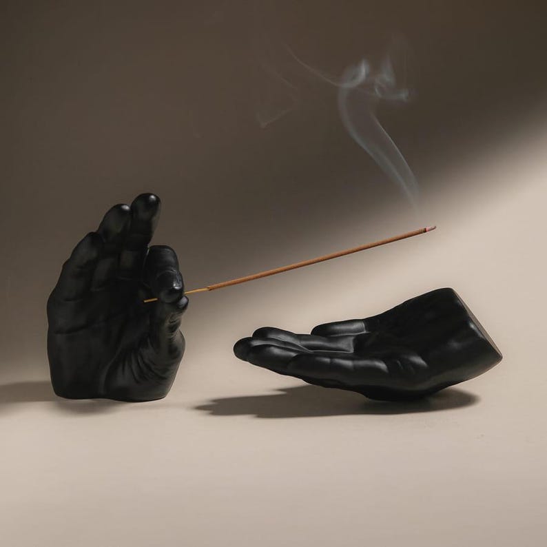May include: A matte black incense holder set consisting of two sculpted hands. One hand is positioned vertically to hold a thin brown incense stick, while the other hand is positioned horizontally with an open palm to catch falling ash.