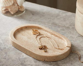 May include: A beige oval tray holds a gold ring and a gold necklace with charms. The tray is made of a textured stone material. A glass jar with seashells is in the background.