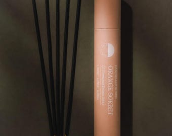 May include: A cylindrical peach-colored cardboard tube labeled Orange Sorbet with five thin black incense sticks arranged in a fan shape. The text on the tube reads Handrolled in India, Orange Sorbet, 20 Ceremonial Incense Sticks, Orange, Cream, Vanilla.