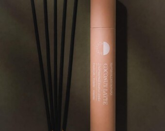 May include: Five thin black incense sticks with yellow tips arranged in a fan shape next to a cylindrical peach-colored cardboard tube. The tube features white text reading Coconut Latte, 20 Ceremonial Incense Sticks, and Handrolled in India.