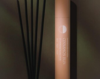 May include: A cylindrical light peach cardboard tube labeled "CINNAMON BUN" with white text, containing 20 ceremonial incense sticks. Five thin black incense sticks with yellow-tipped ends are arranged in a fan shape next to the tube.
