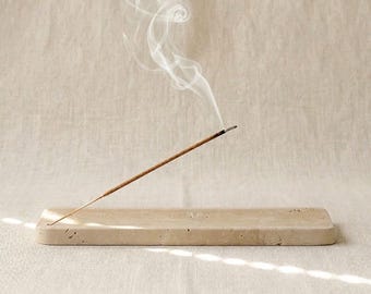 May include: A rectangular beige stone incense holder with a porous, textured surface. A single thin brown incense stick is inserted into the holder at an angle, with a trail of white smoke rising and swirling upward into the air above the object.